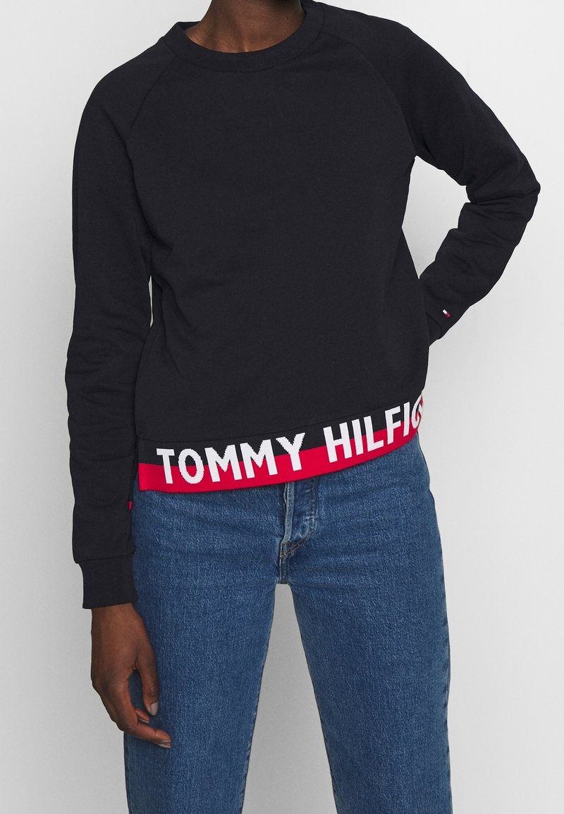 Personne portant un sweat-shirt noir à manches longues avec "Tommy Hilfiger" en lettres blanches sur un liseré rouge et un jean bleu, main sur la hanche.