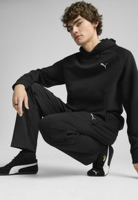 Svart sweatshirt med huva och framfickor, kombinerad med svarta byxor. Båda har subtila grå Puma-logotyper. Svartvita sneakers.