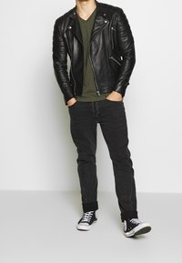 Schwarze Leder-Motorradjacke mit gesteppter Schulterpartie, olivgrünes V-Ausschnitt-Shirt, schwarze Jeans und schwarze hochgeschnittene Sneaker.