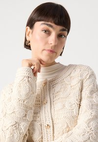 Cremfarbene Häkeljacke mit strukturierten Blumenmustern und goldenen Knöpfen, kombiniert mit einem hellbeigen Rollkragenpullover.