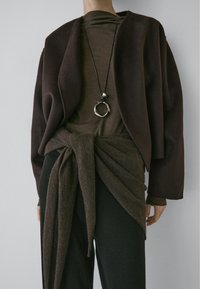 Brown oversized jacket made of soft fabric, layered over a brown top. Features a tied detail at the waist and a silver circular necklace accent.
