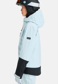 Jasnoniebieska wodoodporna kurtka narciarska z czarną dolną częścią, z przymocowanym kapturem i logo Roxy na rękawie. Biały kask dopełnia całość.