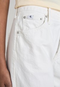 Pantalones cortos de mezclilla blancos con un acabado texturizado, que cuentan con un bolsillo lateral, herrajes metálicos y una etiqueta con el logo de CK en la cinturilla.