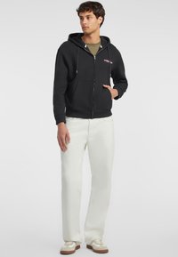 Felpa nera con zip e tasca a marsupio, dotata di cordini. Indossata sopra una camicia khaki, abbinata a pantaloni chiari e sneakers.