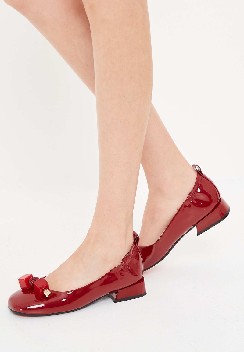Bailarinas de charol rojo con un pequeño tacón, que presentan un detalle de lazo y acentos en contraste en la parte frontal. Textura lisa y diseño minimalista.