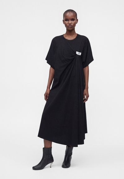 Femme portant une robe midi noire ample avec un détail froncé près de la poitrine et des bottines noires à talons transparents.