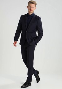 Blond mees, kes kõnnib, seljas tume ehk laevade sinine ülikond koos sobiva särgiga ja mustad nahast kingad, lihtsa heleda tausta ees.