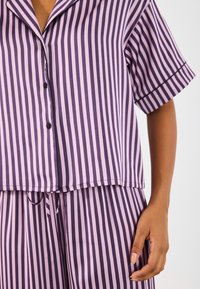 Pruhovaný pyžamový set v levanduľovej a tmavo fialovej farbe, s krátkymi rukávmi, zapínaním na gombíky vpredu a nastaviteľným pásom pre uvoľnený strih.