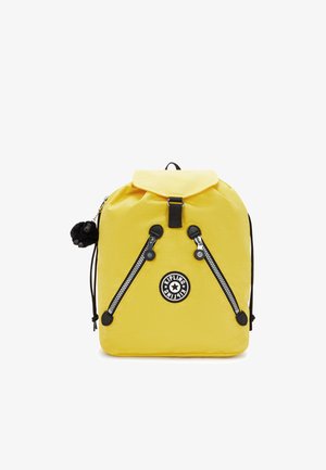 Sac à dos en nylon jaune avec un rabat supérieur, des accents de fermeture éclair noirs, un patch logo circulaire et un porte-clés en peluche en forme de singe attaché.
