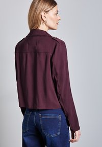 Corta giacca viola con una vestibilità su misura, bottoni decorativi sulle spalle e maniche lunghe, abbinata a jeans di denim blu.