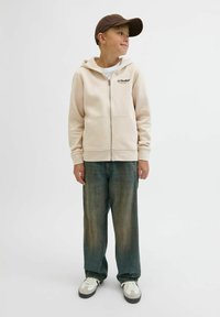 Beżowy hoodie na zamek z białą podszewką i nadrukowanym logo, w połączeniu z szerokimi niebieskimi dżinsami i białymi trampkami z szarymi akcentami.
