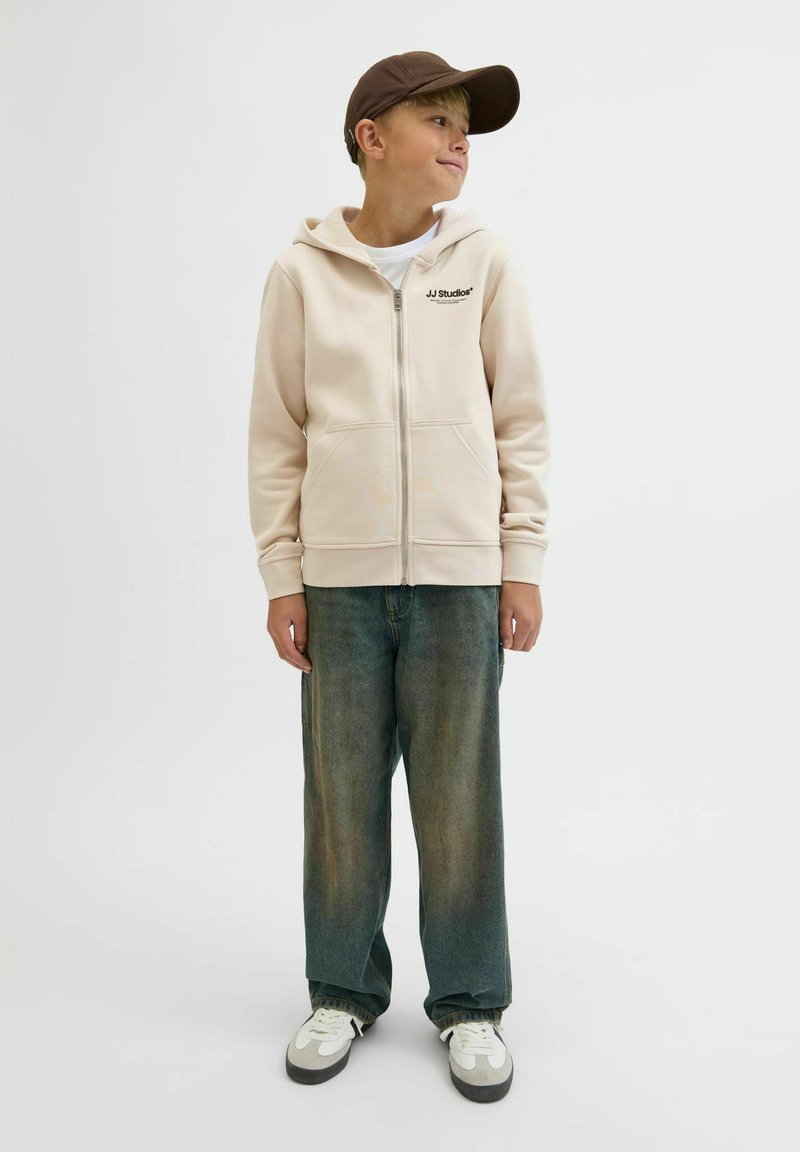 Beżowy hoodie na zamek z białą podszewk�ą i nadrukowanym logo, w połączeniu z szerokimi niebieskimi dżinsami i białymi trampkami z szarymi akcentami.