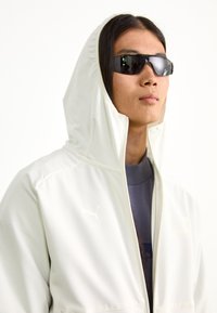 Sudadera blanca de Puma con cremallera completa, tejido elástico y textura suave. Presenta un logo dorado en el pecho y una capucha. El modelo lleva gafas de sol oscuras.