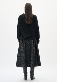 Frau mit langen glatten Haaren, die einen schwarzen, kuscheligen Pullover, einen schwarzen Midirock mit Leopardenmuster und schwarze kniehohe Stiefel trägt, steht mit dem Rücken zur Kamera.