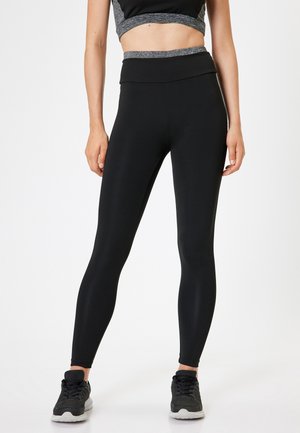 Zwarte high-waisted leggings van rekbare stof, met een gladde textuur en een aangesloten ontwerp. Gecombineerd met zwarte sportschoenen.