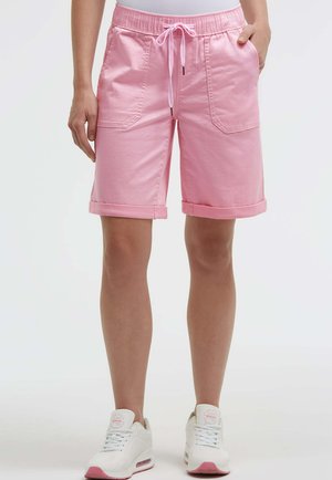 Person trägt rosa Shorts mit Kordelzug und Fronttaschen sowie weiße Sneakers mit rosa Sohlen, steht vor einem schlichten Hintergrund.