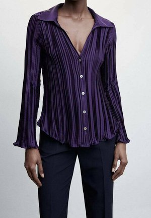 Femme portant un chemisier à manches longues d'un violet profond, plissé verticalement, avec des boutons et un large col, associé à un pantalon foncé.