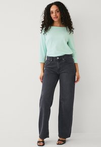 Una modella indossa una camicia a maniche lunghe verde menta e jeans svasati scuri con tasche anteriori, abbinati a sandali neri senza tacco.