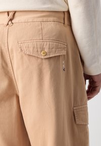 Light brown pants with a back pocket, featuring a button closure and decorative stitching in blue and white. Soft fabric texture.