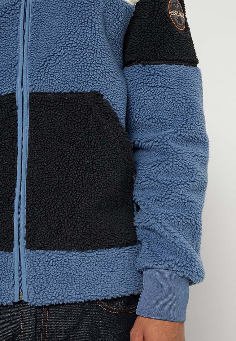 Chaqueta de forro polar en azul y negro, con cremallera frontal, tejido texturizado y puños acanalados. Un parche con el logo en la parte superior de la manga añade un detalle.