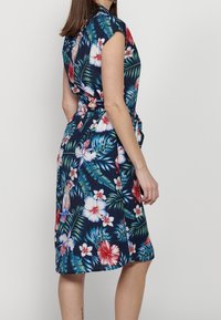 Blumenkleid mit navy Hintergrund, lebhaften Hibiskus- und Palm-Mustern, kurzen Ärmeln und einer Taillierung mit Bindegürtel.