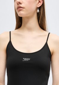 Γυναίκα που φοράει μαύρο τοπ με λεπτές ράντες και λογότυπο "tommy jeans" και ένα ασημένιο σκουλαρίκι σε σχήμα δάκρυ στην αριστερή της αυτί.