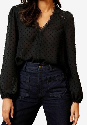 Woman wearing sheer black textured blouse with lace trim and puffed long sleeves tucked into high-waisted dark blue jeans with front pockets.