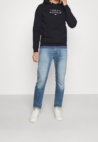 Marineblaue Kapuzenjacke mit "TOMMY HILFIGER"-Logo, Fronttasche, kombiniert mit hellblauen Jeans und weißen Sneakers. Lässiges Outfit vor neutralem Hintergrund.