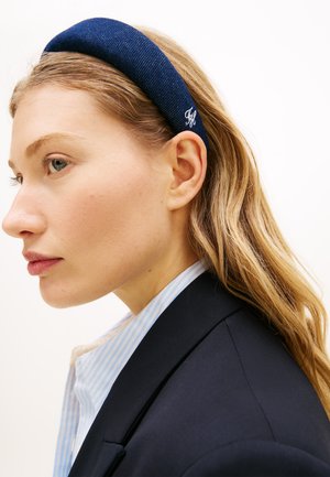 Femme blonde portant un blazer bleu marine et une chemise rayée bleue, coiffée d'un serre-tête bleu marine matelassé avec une petite broderie blanche.