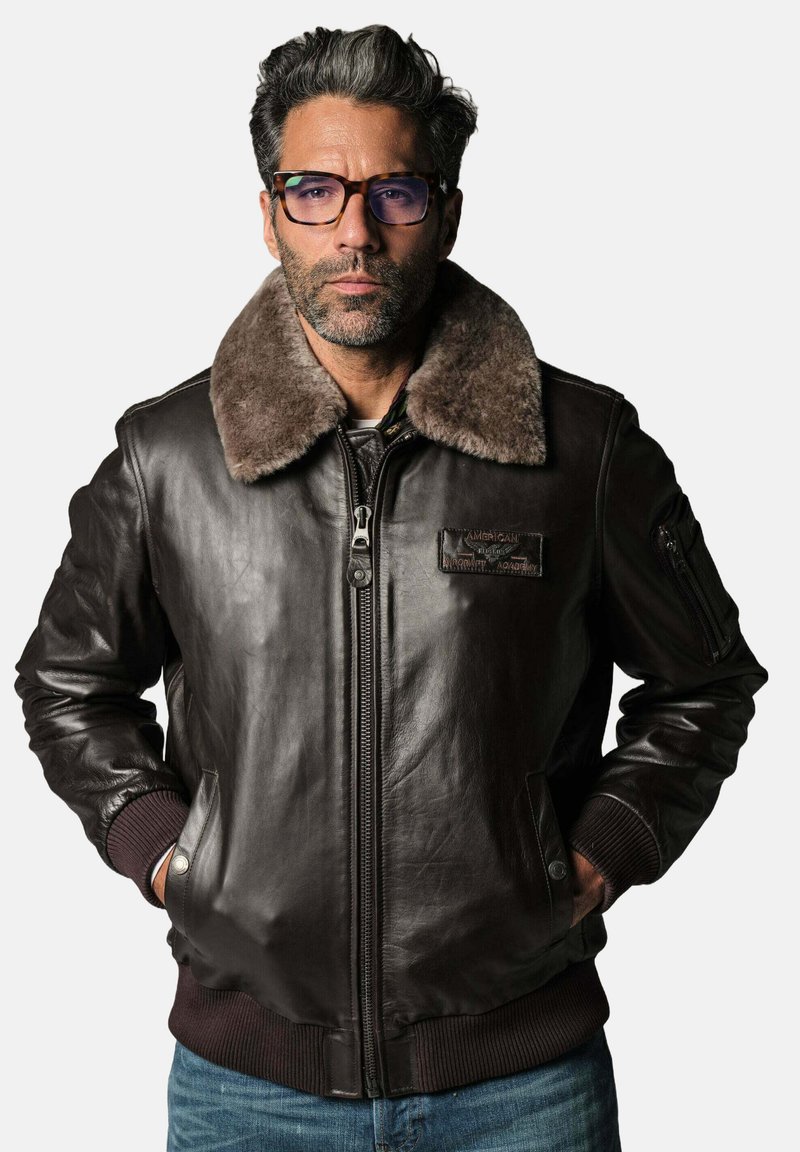 Blouson en cuir marron avec un col en fausse fourrure, des poignets côtelés, une fermeture éclair à l'avant et des poches latérales. Il présente un patch sur la poitrine gauche.