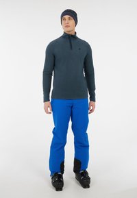 Blå skibukser og en mørk langærmet skjorte med halvlynlås. En sort hue med blå detaljer. Stærke skistøvler fuldender outfitet.