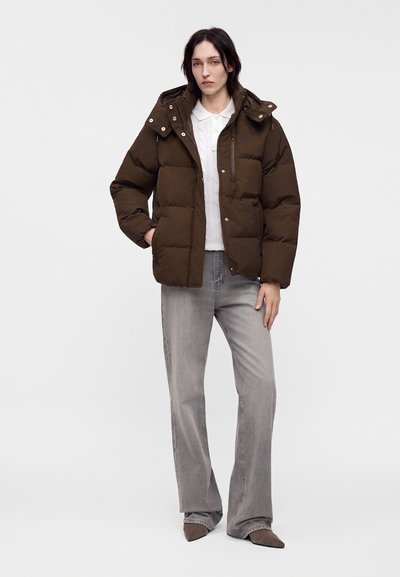 Femme portant une doudoune marron sur une chemise blanche à col, jean gris à jambes larges et chaussures marron pointues, debout devant un fond uni.