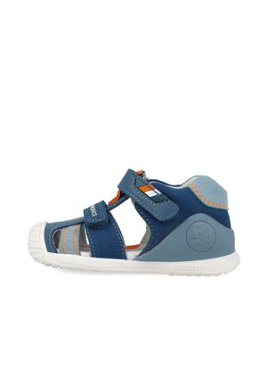 Sandalia infantil azul y gris con costuras naranjas, correas ajustables de velcro y suela blanca flexible.