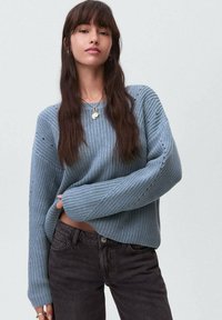 Světle modrý pletený svetr s dlouhými rukávy, ribkovanou texturou a otevřenými stehy, kombinovaný s tmavými džíny. Viditelný kulatý přívěsek na náhrdelníku.