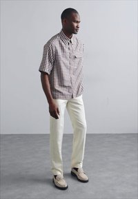 Camisa de manga corta a cuadros en color beige y marrón, combinada con pantalones crema y mocasines en tono marrón claro. Fondo gris con textura suave.