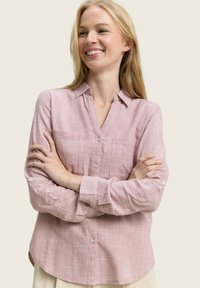 Smiling woman with long blonde hair wearing a light pink checkered button-up shirt with sleeves rolled up, arms crossed, beige background.