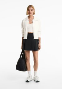 White zip-up sweater, beige cropped top, black pleated skirt, paired with white sneakers and black tote bag. Smooth textures and simple design.