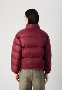 Giacca puffer bordeaux con colletto alto e design trapuntato. Presenta polsini elastici e orlo corto. Indossata con pantaloni in denim chiaro.