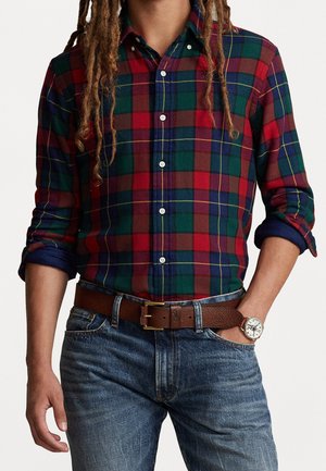 Muž ve sklopené červené, zelené a modré kostkované košili s vyhrnutými rukávy, hnědým koženým pásem, modrých džínách a hodinkách s bílým ciferníkem.