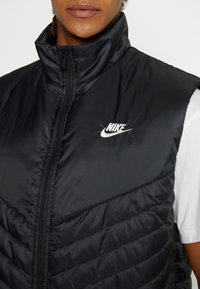 Svart vadderad väst i nylon, med hög krage, dragkedja och vit Nike-logotyp på bröstet. Utmärkande chevronmönster.