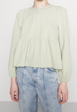 Persoon die een lichtgroene, getextureerde blouse met lange mouwen en een geplooide taille draagt, samen met lichtblauwe jeans met hoge taille.