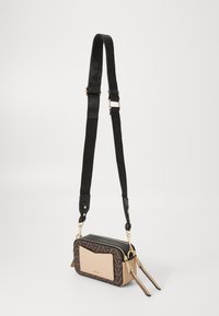 Sac bandoulière avec un motif géométrique marron et noir, un panneau avant lisse couleur beige, des accessoires en or et une sangle réglable.