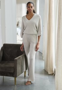 Lichtgrijze geribde gebreide lounge-set met een V-hals top en wijde broek. Zachte textuur, met twee voorzakken. Fluwelen fauteuil in de buurt.