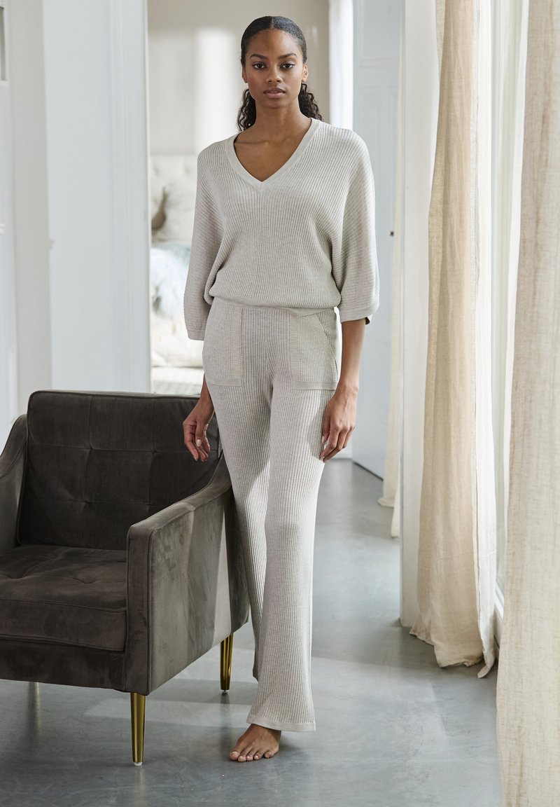 Lichtgrijze geribde gebreide lounge-set met een V-hals top en wijde broek. Zachte textuur, met twee voorzakken. Fluwelen fauteuil in de buurt.