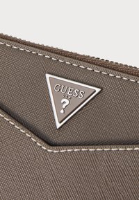 Portefeuille en matériau texturé brun avec des coutures blanches et un logo en argent en forme de triangle portant "GUESS USA". Fermeture zippée sur le dessus.