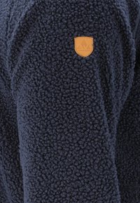 Navy sininen fleece-takki, jossa on teksturoitu kangas, ja ylähihoissa tanssia väriä olevat nahka-logopatchit. Pehmeä ja lämmin muotoilu.