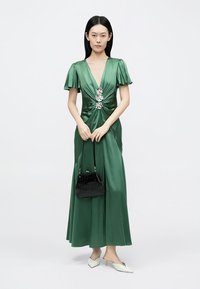 Green satin maxi dress with short flutter sleeves, a deep v-neck, twisted front detail, and silver floral accents. Model holds a black handbag.