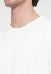 Fehér texturált pulóver kerek nyakkivágással és finoman kidomborodó "BOSS" logóval a mellrészen. Puha anyag, laza szabás.