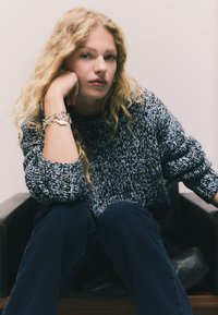 Suéter de punto en tonos azules y grises, con un ajuste holgado y puños acanalados. El modelo lleva una pulsera de plata con múltiples dijes y está sentado en una silla negra.