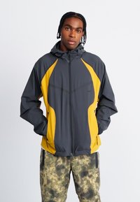 Vindjakke med grå krop og gule detaljer. Har hætte, lynlås og et sporty design. Kombineret med bukser i camouflage-mønster.